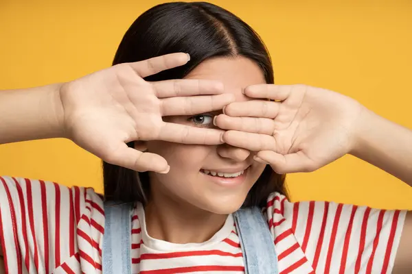 stock image Portrait playful smiling teen girl hiding looking through hand one eye on yellow background. Brunette happy teenager playing hide and seek game. Banner, poster for advertisement, marketing concept.