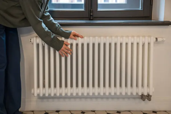 Woman warming hands near radiator at home after walking in cold winter weather, female touching barely warm battery during heating season, person near window checking heating system