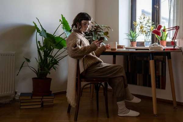 Diligent teenager resting while doing homework, drinking coffee, reading news in internet, scrolling social networks, messaging in smartphone. Teen girl sitting at desk in cozy room with houseplants