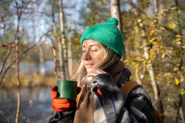 Smiling of pleasure tourist woman standing in wood with cup of hot tea, relaxing after hiking in sun rays with eyes closed, breathing fresh air, listening to birds singing, enjoying pastime in nature