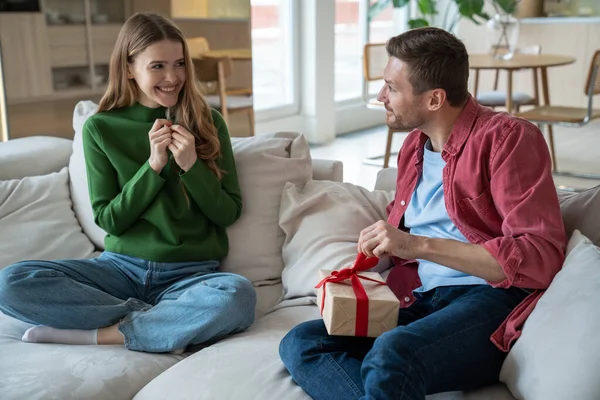 Joyful wife looks at husband unpacking present gift box smiling expecting reaction sit on couch at home. Woman congratulating man. Happy couple celebrating birthday anniversary family holiday together