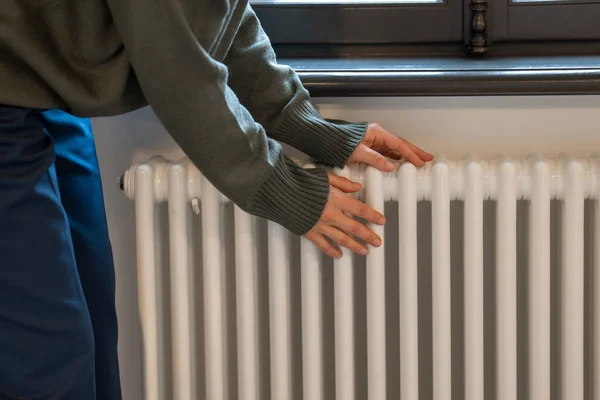 Woman warming hands near radiator at home after walking in cold winter weather, female touching barely warm battery during heating season, person near window checking heating system