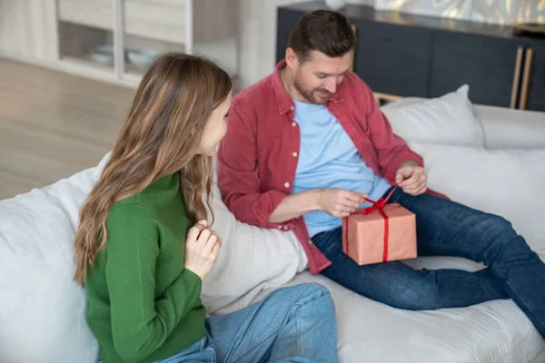 Romantic surprise. Contented man unpacking present gift box with woman look at reaction sit on home couch. Wife congratulating husband. Happy couple celebrate birthday anniversary family holiday.