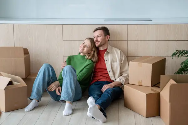 Happy couple man woman hugging dreaming about living in new apartment sitting on floor on kitchen at home. Relocation, moving to flat, family mortgage concept. Spouses enjoying life changes resting.