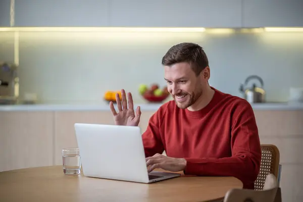 Cheerful man has online business meeting video call sitting on kitchen at home waving hand greeting colleagues looking at laptop screen. Freelance remote distant work job, digital nomad chat concept