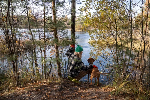 stock image Woman petting dog sitting on haunches in nature autumn park looking at sphagnum swamp, lake. Tourist female travelling with pet friend on weekend sit on haunches, back view. Travel tourism wanderlust.