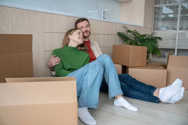 Family couple relaxed man woman dreaming about life in new apartment sitting on floor of modern kitchen. Relocation, moving to flat, family mortgage concept. Happy couple spouses enjoying life changes
