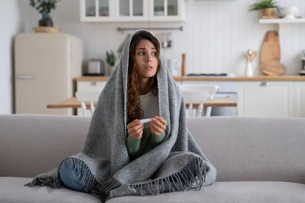 Sick exhausted woman with thermometer to measure body temperature sit on sofa at home wrapped in blanket. Suffering female experiences health problems due to abnormally cold winter weather
