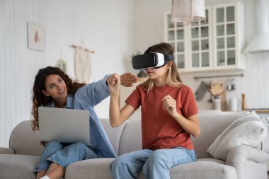 Kadın anne, çocuğun VR 'da bu kadar zaman geçirmesine izin vermiyor. VR kulaklıklı genç kız, kanepeye oturup sanal oyun dünyasını genişletirken annesi yanında dizüstü bilgisayarla çalışıyor. Sanal gerçeklik ve çocuklar