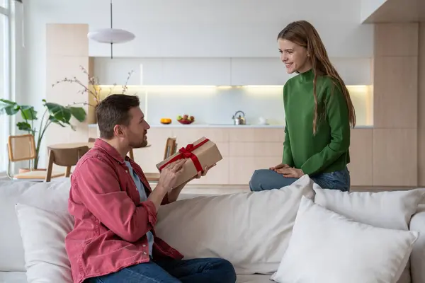 Shiny smiling European woman giving unexpected surprise for birthday, special event, looking with joy at beloved boyfriend, waiting for man reaction to presented cardboard giftbox with red ribbon