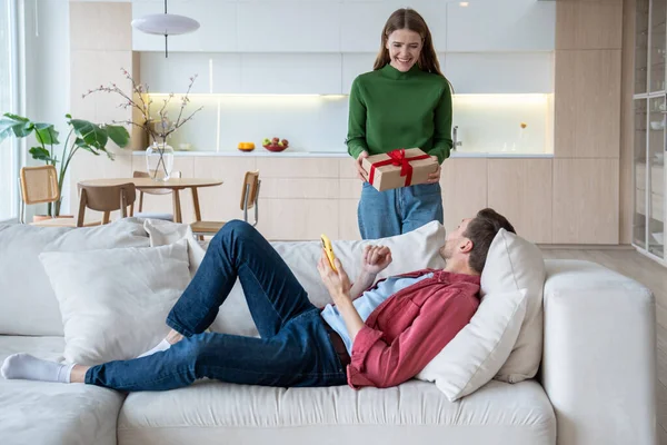 Loving woman presenting gift in cardboard box bound with ribbon to loved man lying on sofa. Male receiving present happy with unexpected surprise. Love, birthday. Tender relations, little family joys