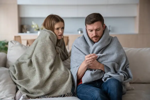 Family couple young man woman measuring temperature sitting on couch wrapping in plaids. Sad frustrated wife husband suffering from seasonal flu or cold. Worry about feeling unhealthy with influenza.