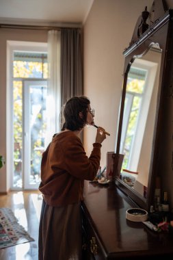 Teenage girl looking in mirror applying everyday makeup on face with brush. Woman standing in room using compact powder to cover flaws. Morning routine before work, putting on make-up before going out clipart