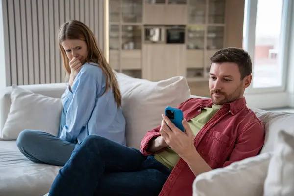Cheating husband messaging, chatting in smartphone, scrolling social networks with happy face expression. Offended upset wife with reproachful glance suspecting of unfaithfulness. Family crisis