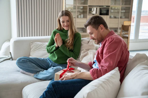 Surprised husband unpacking gift wife waiting sit on sofa near with pray hands looking reaction. Pleased woman congratulate man. Happy couple celebrate birthday, anniversary family holiday together