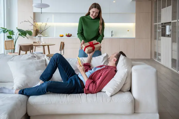 Loving wife presenting gift in cardboard box bound with ribbon to loved husband lying on sofa. Man receiving present happy with unexpected surprise. Birthday. Tender relations, little family joys