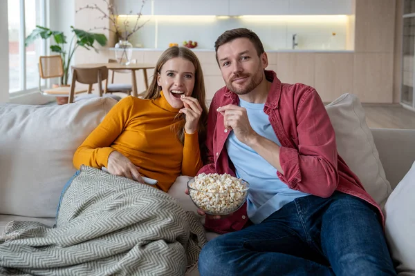 stock image Intrigued couple sitting on sofa, watching favourite humorous show, comedy on TV, eating popcorn, relaxing during weekend. Spending free time, pastime, lockdown, covid isolation together at home