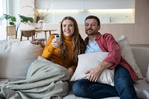 Joyful wife and loving husband enjoying lazy weekends at home. Female covered with warm plaid, holding remote control panel, flipping channels, watching humorous show on TV. Husband resting alongside