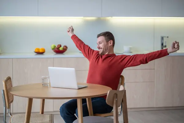 Happy freelancer man stretching arms smiling ending work sitting on kitchen with laptop. Guy having break resting after working on computer at home. Freelance, distant work, job, finishing project.