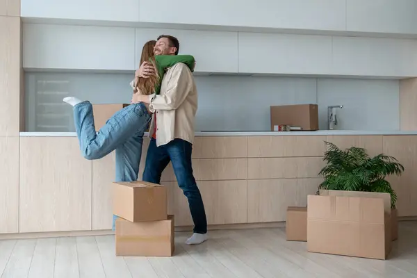 Overjoyed wife and husband hugging on kitchen moving to new flat among cardboard boxes. Relocation, family mortgage, buying apartment, real estate. Happy emotional couple spouses enjoying life changes