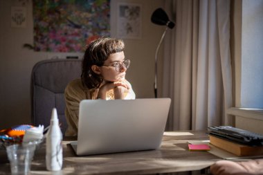 Serbest çalışan bir kadın pencereye bakıp masa başında oturup dizüstü bilgisayarla çalışmaya çalışıyor. Üzgün sıkılmış kız dikkat konsantrasyonu, dikkat eksikliği sendromu, iş motivasyon kaybı yaşıyor..