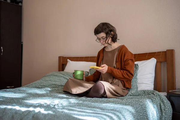 Phone addiction. Thoughtful teen girl reading message scrolling social media in smartphone drinking tea in bed. Morning time of student at home. Focused woman using cellphone, drinks coffee in bed.
