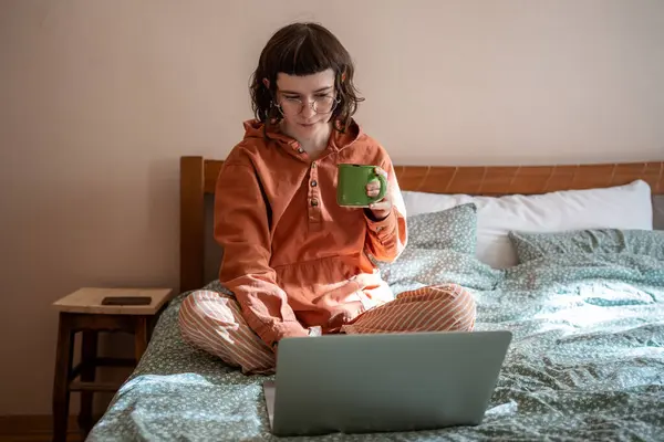 ティーンガールは 自宅で紅茶を飲んでいるベッドに座ってラップトップ上のシリーズ映画を見ています メガネに内向的な女性 孤独で週末にリラックスして休むパジャマ — ストック写真