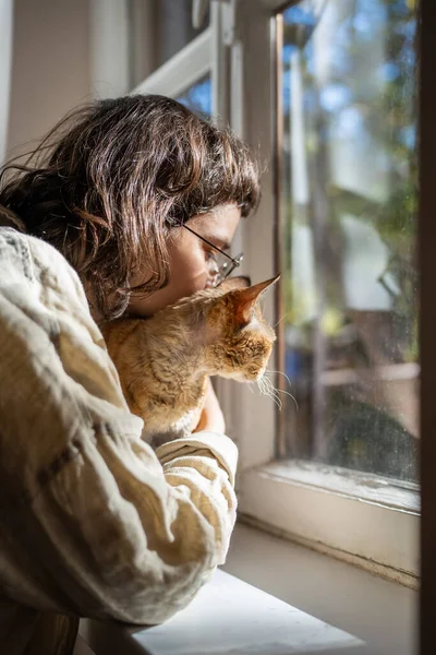 Loving pet owner looking at window with beloved breed cat Devon Rex. Teenager hugging, embracing adorable kitten purring of pleasure. Antistress therapy, unconditional love, friendship with pets