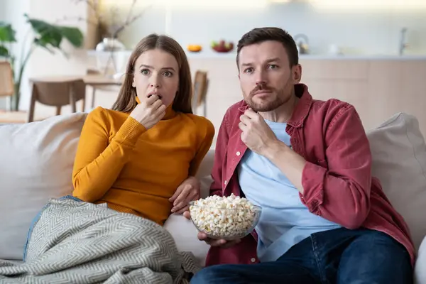 Surprised family couple rest watch TV show, movie, series sitting on couch at home eating popcorn. Amazed relaxed married man and woman enjoy pastime together interested in thriller with exciting plot