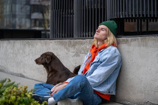 Sad upset stressed hipster guy in depression sitting in city street on ground with sorrow face expression. Homeless stray dog sits aside. Animals giving emotional support in difficult life situations