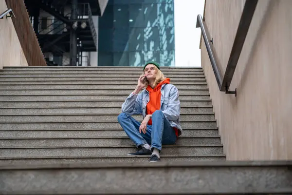 Expressive fashionable skinny male in trendy outwear sitting on staircase, speaking on smartphone. Stylish guy, representative of generation Z looks like tourist, international student, freelancer