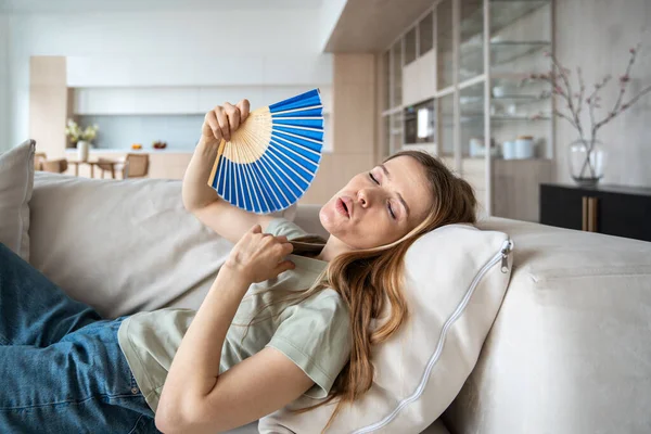 stock image Overheated woman suffering from hot summertime temperature at home. Exhausted female with difficult breath lying on sofa, waving with hand fan in need of cooling ventilator, conditioner, refreshing