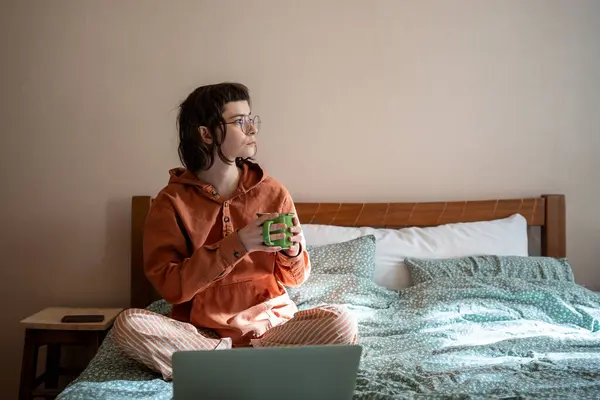 Woman freelancer drinking tea having break sitting in bed at home looking at window. Female in glasses pyjama working. Freelance, remote, distant work job in relaxed atmosphere, self-organization.