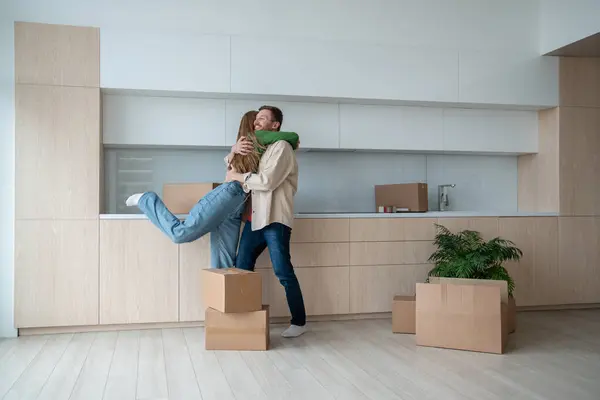 Overjoyed wife and husband hugging on kitchen moving to new flat among cardboard boxes. Relocation, family mortgage, buying apartment, real estate. Happy emotional couple spouses enjoying life changes