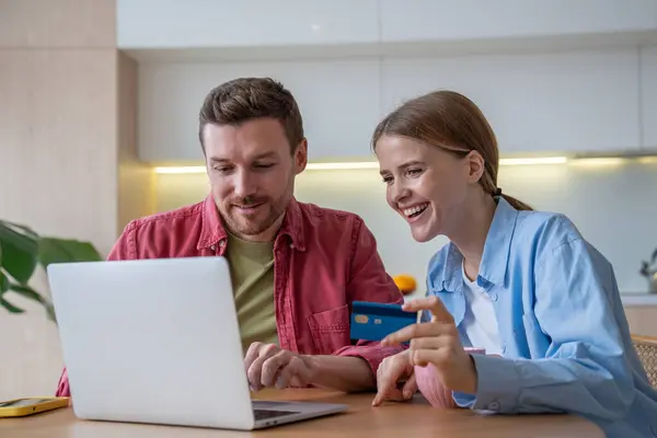 Smiling couple sitting at home, using laptop, surfing web sites, choosing goods in internet stores, buying, purchasing. Cheerful wife and husband spend family budget. Saving time with online shopping