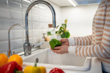 Hands washing green paprika pepper at home. Preparation of fresh salad. Vegetables on worktop near to sink in light kitchen interior. Healthy vegetarian vegan raw food. First step of cooking meals.  clipart