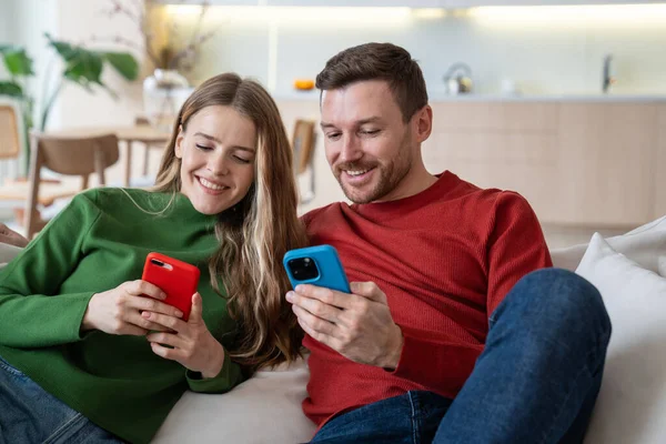 Cheerful happy couple sitting on sofa with smartphones, smiling, playing absorbing online game, watching short funny videos in internet, exchanging photos, having group video call with friends