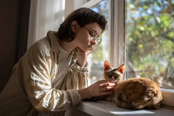 Sad woman feeling lonely, stressed in need of friendship, emotional support, tactile contact, standing near windowsill, caressing, stroking cat Devon Rex, thinking of life. Pet in role of anti stress.