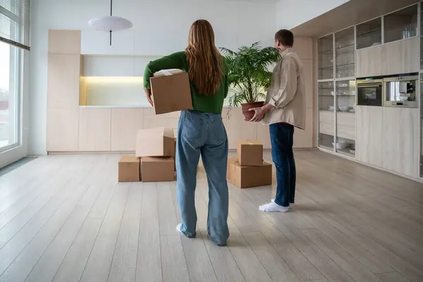 Rear view of new home owners with boxes and houseplant in hands. Family couple taking things to another rented light apartment. Housewarming in mortgage apartment. Beginning of happy married life.