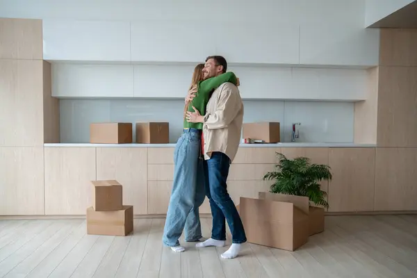 Happy man hugging beloved woman in new apartment. Family couple move together. Wife and husband cuddling enjoy life changing. Satisfied spouses relocating standing with cardboard boxes in new flat.