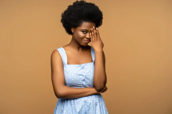 stock image Shy calm bashful african female with natural curly hair, wearing blue summer dress looking into camera but hiding face. Diffident female embarrassed by photographer isolated on beige studio background