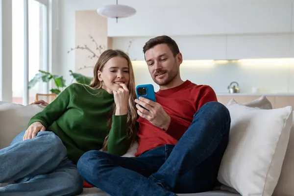 Smiling glad husband sitting on sofa, holding smartphone, scrolling pages. Interested wife looking at screen. Loving couple looking photos, surfing in social networks, watching videos, shopping online