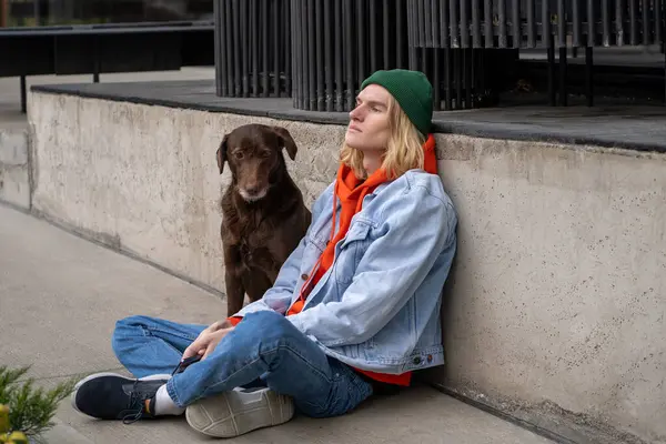 Frustrated upset young man sitting on sidewalk with dog feeling solitude depression have troubles. Unhappy tired hipster guy pet owner spending time outside with dog thinking about difficult breakup.