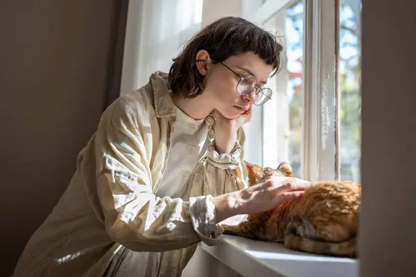Lonely teenage girl need of emotional support, tactile contact standing near window with adorable cat friend. Caring young woman stroking cat sadly looking thinking life troubles. Pet therapy at home.