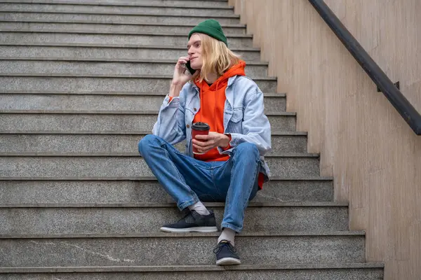 Joyful student guy smiling talking on cellphone sitting on city staircase with takeaway coffee. Contented young hipster man have nice chatting conversation on street with girlfriend on mobile phone.