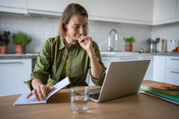 自宅でフリーランスで働く陽気なペンギン女性 テーブルに座って ノートパソコンの画面からテキスト文書を読み コピーブックで論文を書き留める 笑顔の女性は良い仕事の結果に満足しています — ストック写真