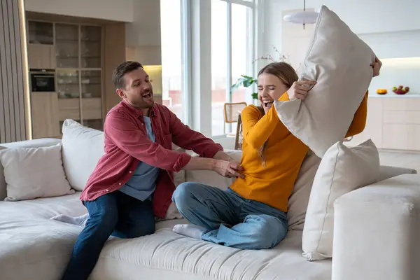 Joyful smiling husband trying to touch laughing wife, fighting against playful man with pillow. Amusement bringing positive feelings, fun, joy, happiness into relations. Enjoying every life moment