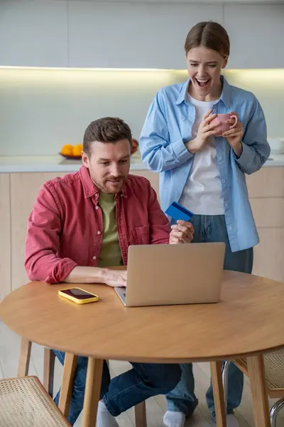 stock image Pleased man sitting kitchen table with laptop and credit card in hands with wife standing next with mug delight with successful online purchase good price. Opportunity to save family budget for couple