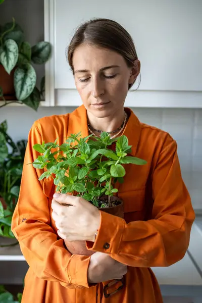 stock image Caring woman tenderly embraces clay pot with home grown mint breathing in sweet aroma. Peaceful female holding potted fragrant herbs looking at leaves. Plant lover standing with home garden result.