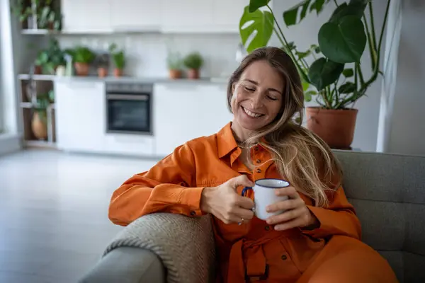 stock image Pleased dreamy middle aged woman sits on couch relax at home holding cup coffee. Joyful female smiling thinking about happy moments, stress free, good mood, wellbeing enjoy weekend time in living room